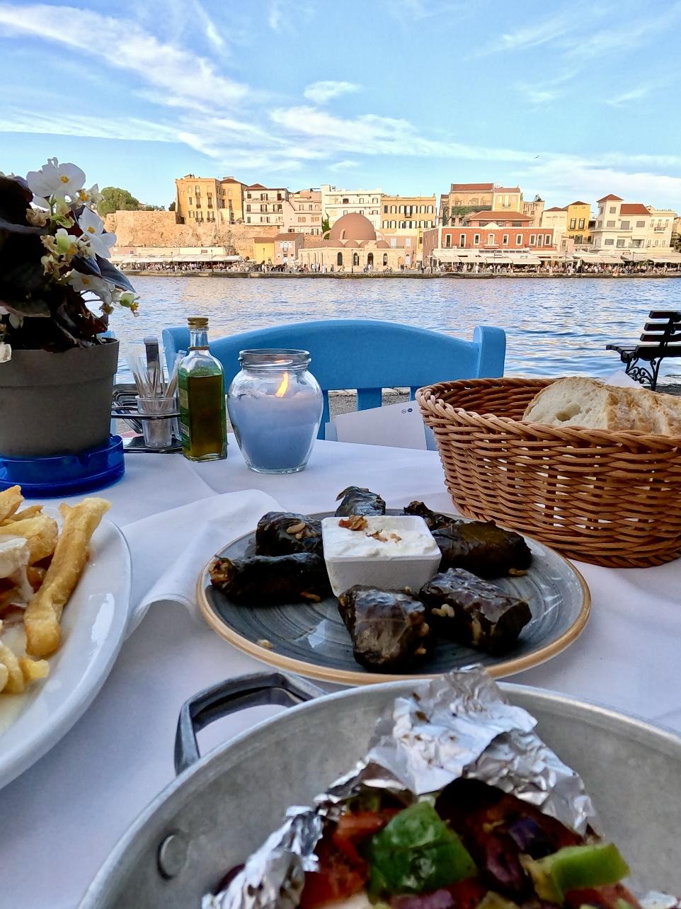 Waterfront dining table with dolmades, Greek salad, and bread overlooking Chania harbor at sunset