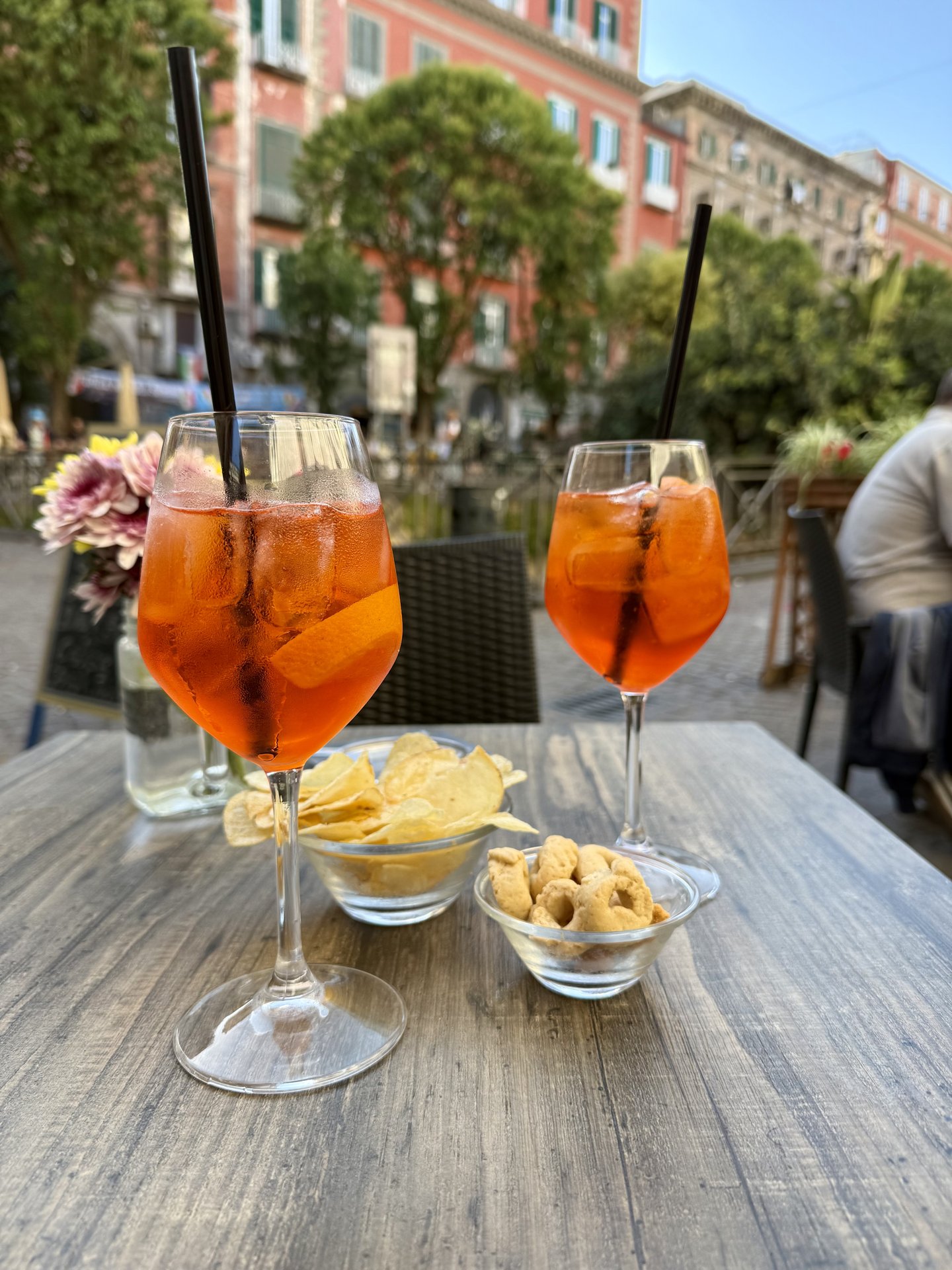 Two Aperol spritzes with snacks at an outdoor café terrace in Naples with colourful buildings in the background