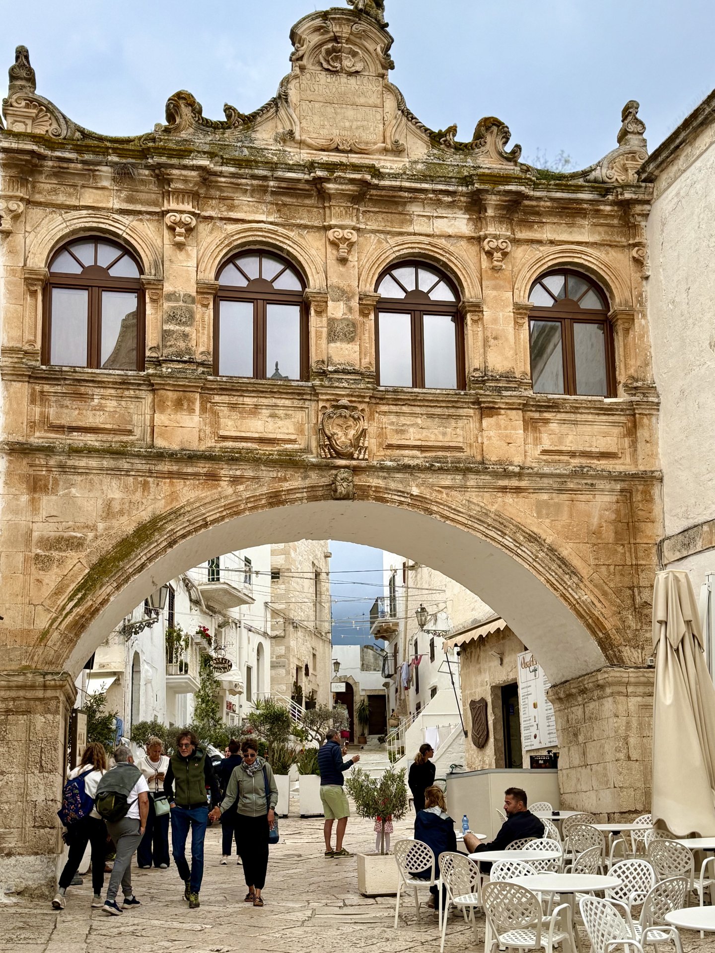 Baroque stone archway in Ostuni's old town with outdoor café tables and visitors walking through